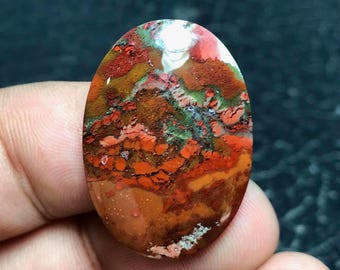 Pode incluir: Gema ovalada com uma vibrante mistura de cores. A pedra apresenta uma combinação de tons vermelhos, laranja, castanhos, verdes e brancos. A superfície polida mostra um padrão único, ideal para joias ou coleção.