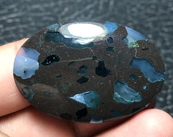 Pode incluir: Gema ovalada com base castanha escura, com manchas azuis, verde-água e pretas. A superfície polida reflete a luz, realçando os padrões naturais. Uma pedra única para joias ou exibição.