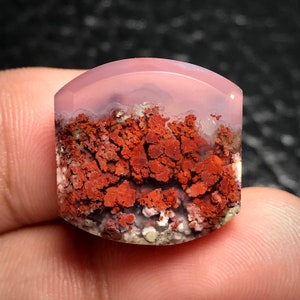 May include: A pink and red agate cabochon with a unique pattern of red and white inclusions. The agate is polished and has a smooth, rounded surface.