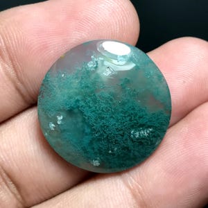 Puede incluir: Una gema redonda y pulida con una base translúcida de color gris pálido. La piedra presenta intrincadas inclusiones en forma de helecho en tonos verde azulado y verde. Un área ovalada blanca y brillante es visible en la superficie superior. La gema se sostiene en una mano.