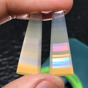 Puede incluir: Dos gemas alargadas en forma de trapecio. Las piedras tienen un degradado de color, que va del amarillo pálido en la base al blanco translúcido en la parte superior. Una piedra muestra bandas iridiscentes.
