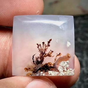 May include: A square-cut dendritic agate gemstone with a translucent, milky white base. The stone features intricate, dark brown, tree-like fern inclusions and patches of metallic, silver-toned mineral deposits embedded within the smooth surface.