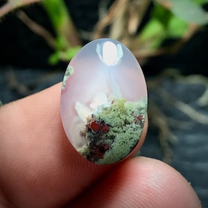 May include: A white agate cabochon with a green and red moss agate inclusion. The agate is translucent and has a smooth, polished surface.