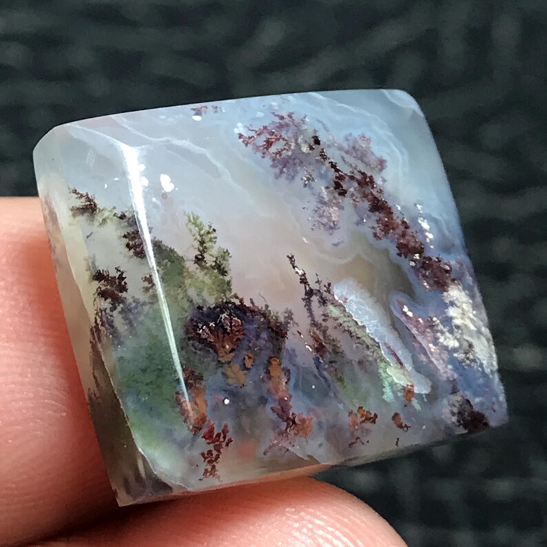 May include: A square-cut gemstone with a translucent, light blue base. The stone features intricate inclusions of brown, green, and white, resembling a miniature landscape. The polished surface reflects light, highlighting the natural patterns within the stone.