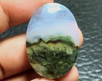 May include: An oval-shaped gemstone with a landscape design. The top portion is a light blue and white, transitioning to a green and brown landscape at the bottom. The stone is held in a person's hand.