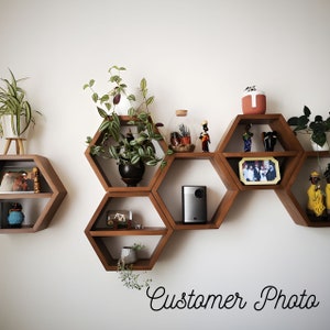 Könnte beinhalten: Ein Satz hexagonaler Holzregale in einem Wabenmuster, an einer weißen Wand montiert. Die Regale sind mit Pflanzen, Figuren, einem Projektor und einem gerahmten Foto dekoriert. Der Text "Customer Photo" ist unten im Bild in Kursivschrift geschrieben.
