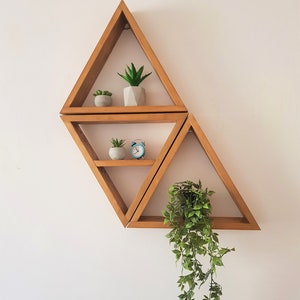 Peut inclure: Trois &eacute;tag&egrave;res triangulaires en bois mont&eacute;es sur un mur blanc. L'&eacute;tag&egrave;re du haut a deux petites plantes en pot, l'&eacute;tag&egrave;re du milieu a une petite plante en pot et un r&eacute;veil bleu, et l'&eacute;tag&egrave;re du bas a une plante suspendue avec des feuilles vertes.