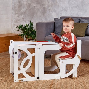 May include: A white wooden children's art table and chair set with a roll of paper and a pencil. The table has a cutout design and the chair has a curved back. A young child is sitting at the table drawing.