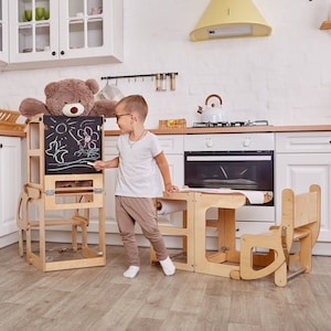 Puede incluir: Una torre de aprendizaje de madera para niños con una pizarra y un rollo de papel. Un niño está dibujando en la pizarra con una tiza amarilla. Un oso de peluche está sentado en el estante superior de la torre. Hay una segunda torre de aprendizaje de madera con una mesa y una silla. Las torres están en una cocina blanca.