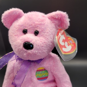 May include: Pink plush Easter bunny with a purple ribbon and an embroidered Easter egg on its chest. The bunny has a black nose and black eyes. The bunny is wearing a Ty Beanie Baby tag.