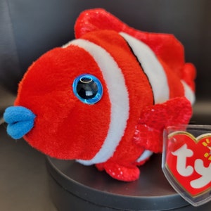 May include: Red and white striped plush clownfish with blue eyes and a blue fin. The fish is sitting on a black surface with a Ty Beanie Baby tag.