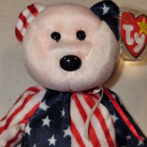 May include: A pink and white beanie baby bear wearing a red, white, and blue American flag outfit. The bear has black eyes and a black nose. The bear has a white tag with the Ty logo and a red heart.
