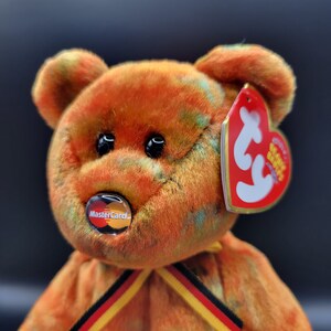 May include: A brown and orange fuzzy teddy bear with a red Ty tag and a Mastercard logo on its chest. The bear is wearing a yellow and black ribbon around its neck.