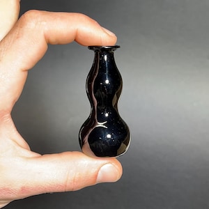 May include: A small, glossy black glass vase with a bulbous body and a narrow neck. The vase is held between fingers against a gray background. The vase has a smooth, reflective surface.