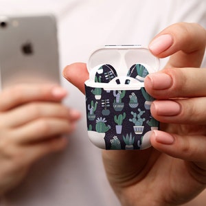 May include: White AirPods case with a dark blue cactus pattern, held open to display the earbuds. The case and earbuds feature a repeating design of various cacti in pots. A silver smartphone is held in the background.