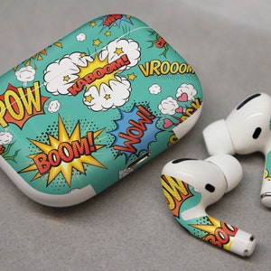 Könnte beinhalten: Weiße kabellose Ohrhörer mit einem türkisfarbenen, gelben und roten Comic-Druckdesign. Das Etui hat die Worte "POW!" "BOOM!" "KABOOM!" "VROOOOM!" und "WOW!" aufgedruckt.