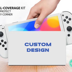 Könnte beinhalten: Weiße Nintendo Switch Konsole mit einer weißen Schutzhülle, auf der "CUSTOM DESIGN" steht. Die Konsole wird von einer Hand gehalten.