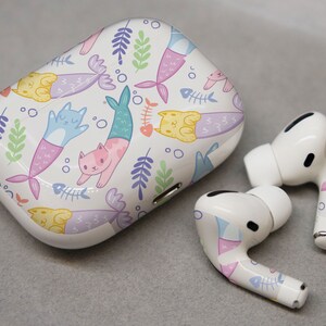 May include: White wireless earbuds with a mermaid and cat design. The case is white with a colorful mermaid and cat design.