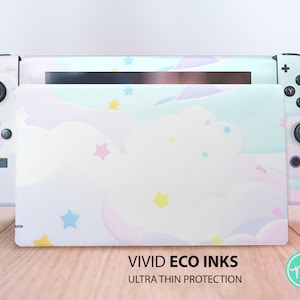 Puede incluir: Una consola Nintendo Switch blanca con una piel colorida, en colores pastel, con nubes y estrellas. La piel está etiquetada con "VIVID ECO INKS ULTRA THIN PROTECTION".