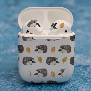 May include: White wireless charging case with a hedgehog and leaf pattern. The case has a hinged lid and is designed to hold wireless earbuds.