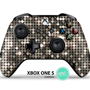 May include: A black Xbox One S controller with a gold and silver sequin pattern. The controller has the standard Xbox buttons, including A, B, X, Y, and a directional pad. The text "XBOX ONE S GAMING" is printed below the controller.