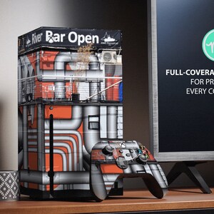 May include: A black and gray video game console with a colorful design featuring a building with pipes and the text "River Bar Open". The console is next to a black and gray video game controller. The text "FULL-COVERAGE KIT FOR PROTECT EVERY CORNER" is visible on a black background.