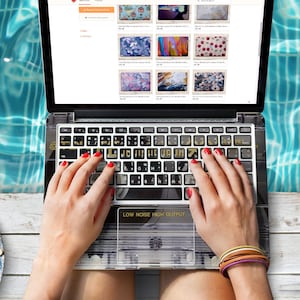 May include: A laptop computer with a custom keyboard cover featuring a cassette tape design. The laptop is open and the screen shows a website with images of different laptop skins. The text "LOW NOISE HIGH OUTPUT" is visible on the keyboard cover.