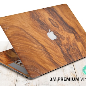 Może przedstawiać: Laptop z wzorem słojów drewna. Laptop ma srebrne logo Apple na pokrywie. Tekst "3M PREMIUM VINYL" jest wydrukowany na dole obrazu.