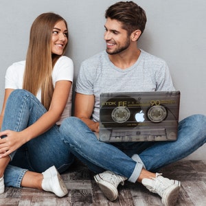 May include: A couple sitting on the floor, the man is holding a laptop with a black and white cassette tape design, the woman is looking at the man, both are wearing jeans and white sneakers.