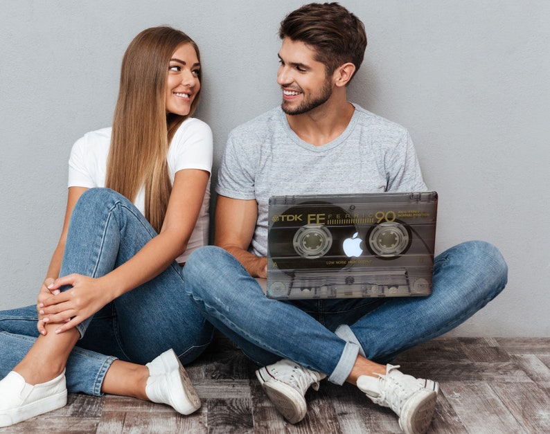 May include: A couple sitting on the floor, the man is holding a laptop with a black and white cassette tape design, the woman is looking at the man, both are wearing jeans and white sneakers.