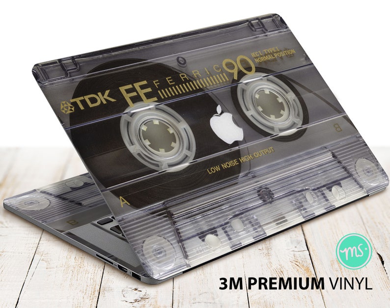 May include: A laptop computer with a clear vinyl skin featuring a vintage cassette tape design. The tape is labeled "TDK FE FERRIC 90" and has the text "LOW NOISE HIGH OUTPUT" printed on it.