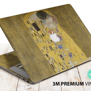 Peut inclure: Un autocollant pour ordinateur portable représentant l'œuvre "Le Baiser" de Gustav Klimt. Le design présente les motifs dorés et en mosaïque du tableau. L'autocollant est appliqué sur un ordinateur portable, avec le logo Apple visible. Le texte "3M PREMIUM VINYL" est imprimé en bas.