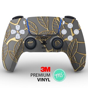Könnte beinhalten: Ein grau-goldener PlayStation 5-Controller mit einem Blatt-Muster. Der Controller hat ein 3M-Logo und den Text "PREMIUM VINYL" unten.