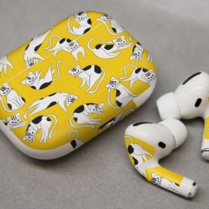 May include: A white wireless charging case with a yellow background and a repeating pattern of black and white cats. The case is designed to fit Apple AirPods Pro. Two white wireless earbuds with a similar cat pattern are also shown.