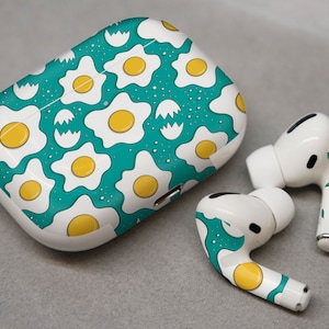 May include: White wireless earbuds with a green and yellow fried egg pattern. The case is also covered in the same pattern.