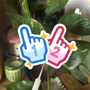 May include: Two foam finger stickers, one blue with a number 1 and the other pink with a number 2. Both have a white outline and yellow starburst accents. The stickers are held by a silver tweezer.