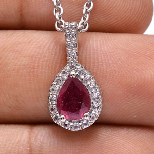 Può includere: Una collana con pendente in argento con una pietra preziosa rosso scuro a forma di pera, circondata da piccole pietre trasparenti. Il pendente è appeso a una delicata catena d'argento. Il gioiello è tenuto in una mano.