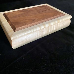 May include: A rectangular wooden box with a light-colored, wavy-grained body and a dark brown, flat top. The top is recessed within a lighter wood frame. The box is closed and sits on a dark surface.