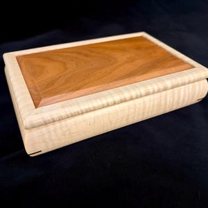 May include: A rectangular wooden box featuring a light-colored, wavy-grained maple wood frame and a smooth, reddish-brown cherry wood lid insert. The box has rounded edges and a beveled top surface, showcasing natural wood grain patterns throughout.