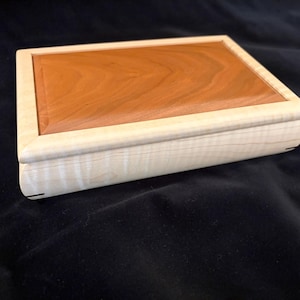 May include: A rectangular wooden box featuring a light-colored, pale wood frame with a visible grain pattern and a contrasting, smooth, reddish-brown wood panel inset on the lid. The box has rounded edges and a clean, finished construction.