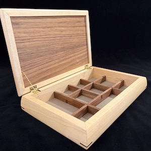 May include: Open wooden jewelry box with a light-colored frame and a darker wood lid. The interior features a grid of small compartments, perfect for organizing rings, earrings, and other small items. The box is hinged with brass hardware.