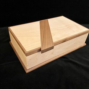 May include: A rectangular wooden box with a light-colored body and a darker wood trim and accent. The lid features a vertical, contrasting wood piece. The box is likely for storage or decorative purposes.