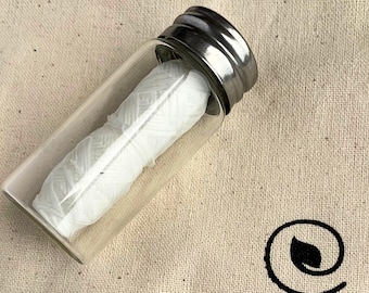 May include: A glass jar with a silver lid containing white dental floss. The jar is sitting on a beige fabric surface.