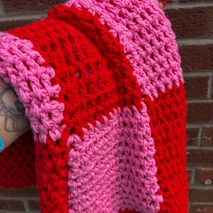Könnte beinhalten: Eine handgefertigte Häkeldecke in einem lebendigen Farbblock-Design. Die Decke hat abwechselnde Abschnitte aus leuchtend roter und hellrosa Wolle. Das dicke Garn erzeugt ein strukturiertes, gemütliches Aussehen. Die Decke ist gefaltet und zeigt das Farbmuster.
