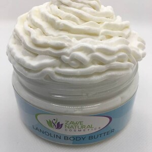 Puede incluir: Un tarro blanco de manteca corporal de lanolina con una textura blanca similar a la crema batida en la parte superior. El tarro tiene una etiqueta azul y blanca con el texto "Zave Natural Cosmetics" y "Lanolin Body Butter".