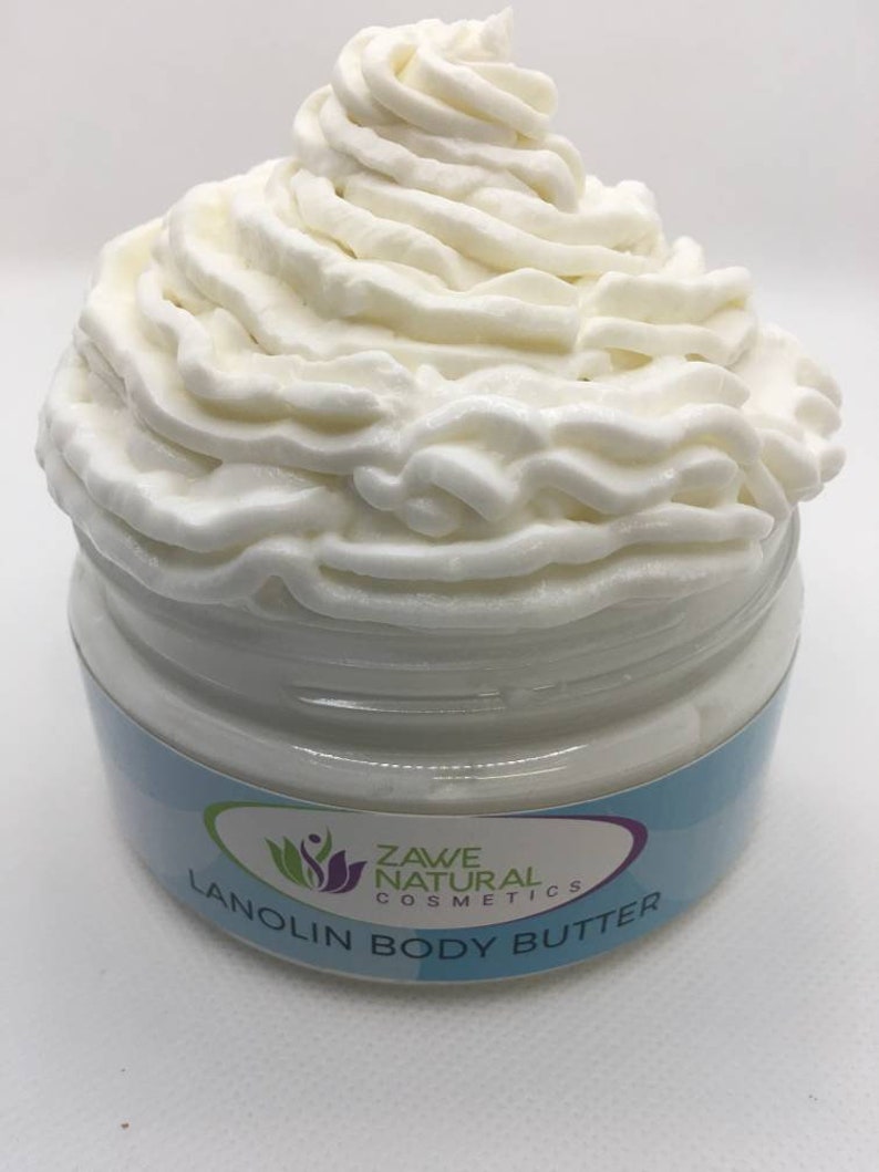 Puede incluir: Un tarro blanco de manteca corporal de lanolina con una textura blanca similar a la crema batida en la parte superior. El tarro tiene una etiqueta azul y blanca con el texto "Zave Natural Cosmetics" y "Lanolin Body Butter".
