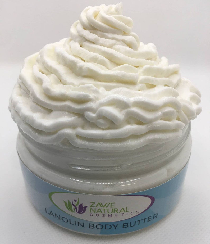 Puede incluir: Un tarro blanco de manteca corporal de lanolina con un remolino de crema blanca en la parte superior. El tarro tiene una etiqueta azul y blanca con el texto "ZAW&Eacute; NATURAL COSMETICS LANOLIN BODY BUTTER".