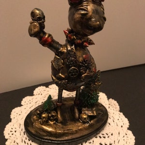 Puede incluir: Una figurita de estilo steampunk en oro y negro de un pájaro con una calavera en la mano. El pájaro tiene detalles rojos y está de pie sobre una base con calaveras y vegetación.