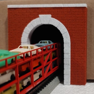 May include: Model train scene featuring a red brick tunnel with a white arched entrance. A train car loaded with miniature cars is emerging from the tunnel. The scene is set against a neutral background.