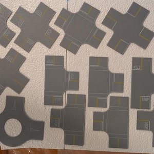 May include: A collection of gray, die-cut road pieces for a model city or train set. The pieces include intersections, T-junctions, and a roundabout, with yellow and white road markings. Some pieces have "STOP" text.